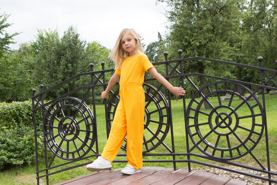 A little girl of six-seven years old with long blond hair in a plain yellow outfit stands and smiles in a summer park on a decorative bridge and poses in full growth.