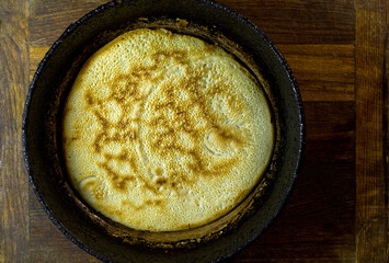 Freshly baked pancake on a black vintage frying pan standing on a wooden board.