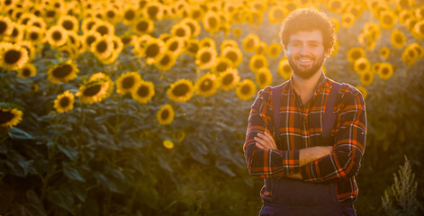 Handsome farmer standing in the middle of a golden sunflower field with his arms crossed and smiling, during a majestic sunrise.