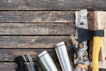 Stainless cup, parts of tent, axe, birch logs and rope on the wooden table background with copy space. Camping.