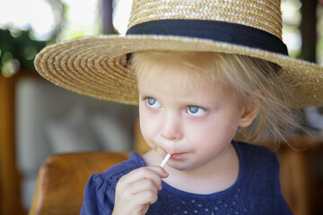 Candid headshot of adorable blonde toddler girl wearing a straw hat and eating candy lollipop	