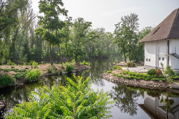 Water around the house with white walls and an authentic roof. Beautiful landscape. Summer time. Ashore green trees