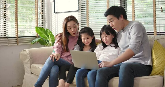 Happy asian family parents and little children enjoy using devices together sit on sofa, technology addicted couple with kids having fun with laptop computer at home. Social distancing self isolation.