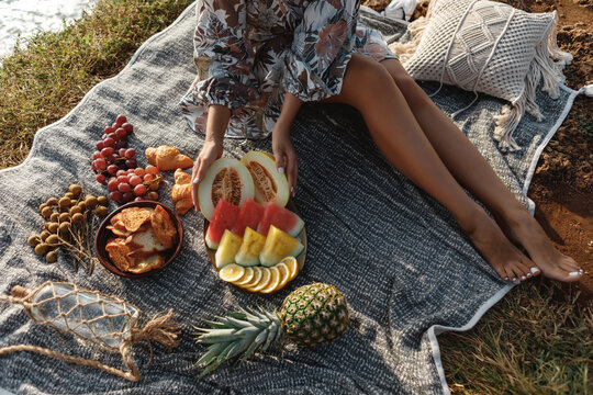 Beautiful Women Seat Near Picnic On The Beach At Sunset In Boho Style. Organic Fresh Fruit On Linen Blanket. Vegetarian Eco Idea For Weekend Picnic