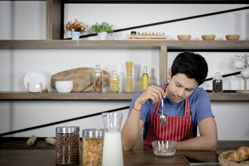 Young Asian handsome man feels stressed and bored while eating, having breakfast meal on wooden table in kitchen room.