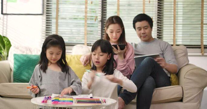 Focused on cute little kids daughters drawing coloring picture while young parents couple relaxing on sofa using device in living room enjoying family leisure hobbies spend free time at home together.