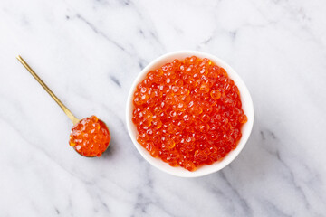 Red salmon caviar in white bowl and golden spoon on marble background. Top view.