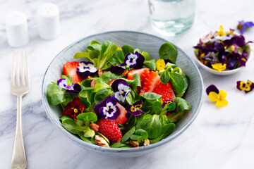 Fresh green salad with strawberries and edible flowers in a bowl. Marble background. Close up.