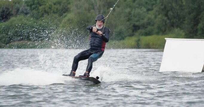 Wakeboarding in a lake near the forest, adult man surfs on the water, ride on a wakeboarding board and performs tricks, jumps on a springboard, 4k 50fps.