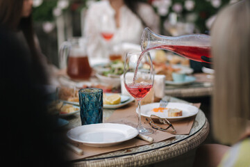 Waiteress pouring rose wine in glass. Background of people eating dinner at summer terrace in cafe. Food concept outdoors. Different kind of food and drinks on the table