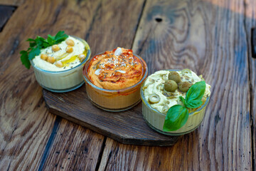 Three types of classic appetizer hummus with paprika and garlic, olives, chickpeas, tahini, olive oil, and oriental spices in a bowl on a wooden table. Traditional Mediterranean vegetarian snack.