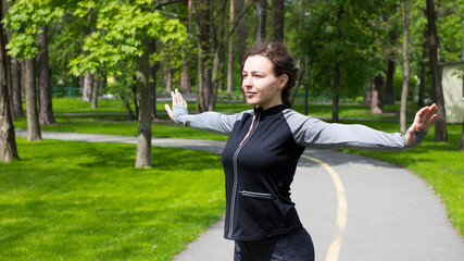 Young woman doing yoga in green city park and looking away.Sporty girl smiling enjoying nature during breathing exercise.Fitness female stretching arms and looking away outdoor.Healthy body lifestyle