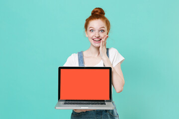 Excited readhead girl in casual denim clothes isolated on blue turquoise background. People lifestyle concept. Mock up copy space. Hold laptop pc computer with blank empty screen put hand on cheek.