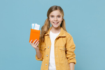 Smiling little blonde kid girl 12-13 years old in yellow jacket isolated on blue background. Passenger traveling abroad to travel on weekends getaway. Air flight journey concept. Hold passport ticket.