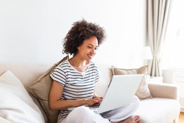 Young beautiful woman with curly hair working on laptop computer while sitting on the sofa at home,...