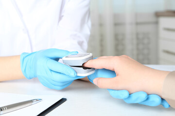 Doctor examining patient with fingertip pulse oximeter at white table in office, closeup