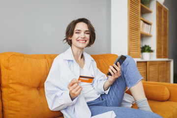 Charming young woman in casual clothes sit on couch spending time in living room at home. Rest relax good mood leisure lifestyle concept. Mock up copy space. Using mobile phone, hold credit bank card.