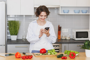 Smiling young housewife woman girl in white casual clothes using mobile phone looking at recipe preparing vegetable salad cooking food in light kitchen at home. Dieting healthy lifestyle concept.