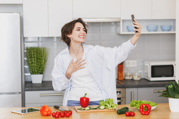 Cheerful housewife woman in casual clothes preparing vegetable salad cooking food in light kitchen at home. Dieting healthy lifestyle concept. Doing selfie shot on mobile phone, put hand on chest.