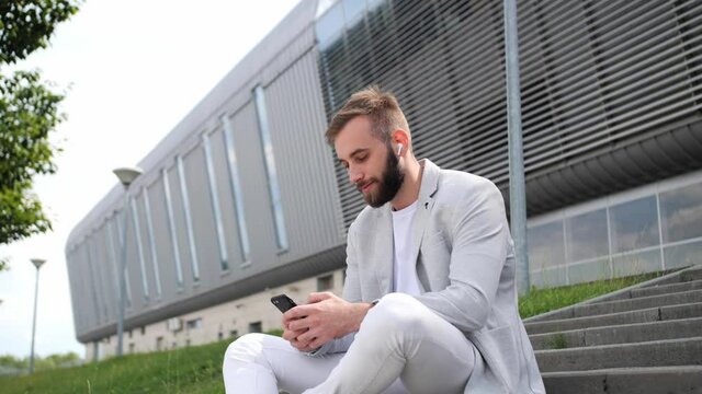 Handsome young man with a beard listens audio book through wireless headphones using a mobile phone, application background modern building light suit internet.