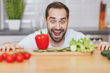 Surprised excited young bearded man guy in white casual t-shirt posing preparing vegetable salad cooking food in light kitchen at home. Dieting healthy lifestyle concept. Mock up copy space.