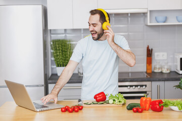Funny young bearded man in casual t-shirt with headphones using laptop computer looking at recipe...