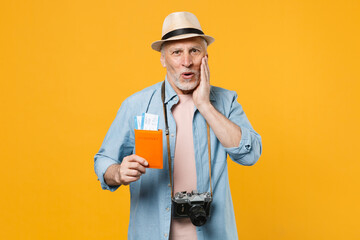 Amazed traveler tourist elderly gray-haired man in hat photo camera isolated on yellow background. Passenger traveling abroad on weekends. Air flight journey. Hold passport tickets, put hand on cheek.