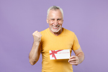 Joyful elderly gray-haired mustache bearded man in yellow t-shirt posing isolated on violet wall background studio portrait. People lifestyle concept. Hold gift certificate doing winner gesture.