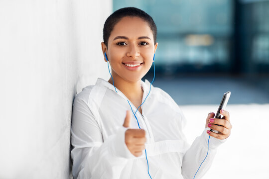 fitness, sport and technology concept - happy smiling young african american woman with earphones listening to music on smartphone and showing thumbs up outdoors
