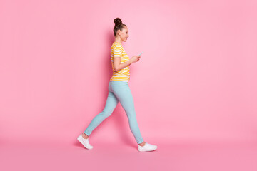 Fototapeta na wymiar Full length profile photo of attractive lady two funny buns hold telephone hands writing new blog post walk down street wear casual striped t-shirt pants shoes isolated pink color background