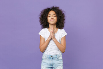 Pleading little african american kid girl 12-13 years old in white t-shirt isolated on violet wall background studio. Childhood lifestyle concept. Mock up copy space. Holding hands folded in prayer.