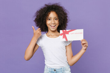 Cheerful little african american kid girl 12-13 years old in white t-shirt isolated on violet wall background. Childhood lifestyle concept. Mock up copy space. Hold gift certificate showing thumb up.