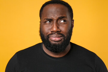 Close up of puzzled bewildered african american man football fan in casual black t-shirt isolated on yellow background studio portrait. People lifestyle concept. Mock up copy space. Looking aside.