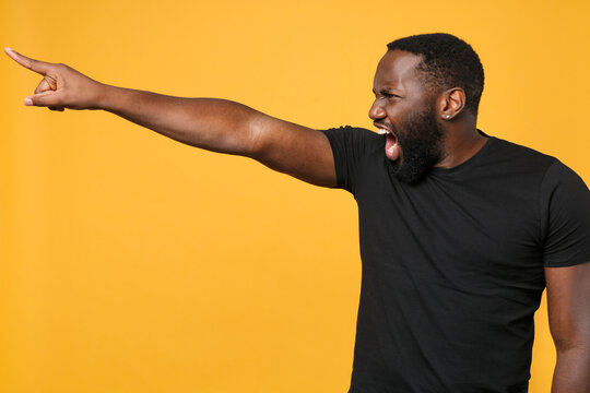 Angry african american man football fan in casual black t-shirt isolated on yellow wall background studio. People lifestyle concept. Mock up copy space. Pointing index finger aside screaming swearing.