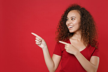 Smiling young african american woman girl in casual t-shirt posing isolated on red background studio portrait. People emotions lifestyle concept. Mock up copy space. Pointing index fingers aside up.