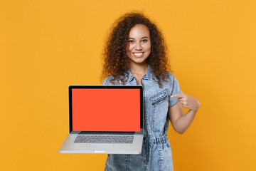 Naklejka premium Smiling young american girl in casual denim clothes isolated on yellow background. People lifestyle concept. Mock up copy space. Pointing index finger on laptop pc computer with blank empty screen.
