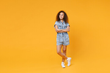 Full length portrait of smiling young african american girl in denim clothes isolated on yellow background studio. People lifestyle concept. Mock up copy space. Holding hands crossed looking aside.