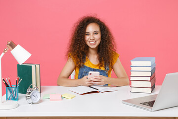 Smiling african american girl employee in office sit isolated on pink background. Achievement business career. Education in school university college concept. Using mobile phone, typing sms message.