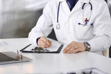 Unknown male doctor sitting and working with clipboard of medication history record in clinic at his working place, close-up. Young physician at work. Perfect medical service, medicine concept