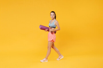 Full length portrait of smiling young fitness woman girl in sportswear working out isolated on yellow background studio. Workout sport motivation lifestyle concept. Hold yoga mat, bottle of water.
