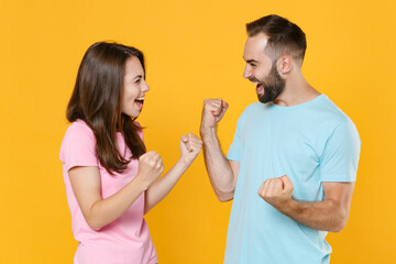 Happy young couple two friends guy girl in blue pink t-shirts isolated on yellow background studio. People lifestyle concept. Mock up copy space. Clenching fists like winner, looking at each other.