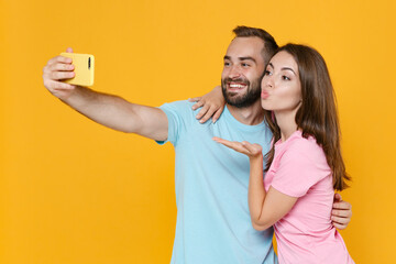 Funny couple two friends guy girl in blue pink t-shirts isolated on yellow background. People lifestyle concept. Mock up copy space. Doing selfie shot on mobile phone hugging blowing sending air kiss.