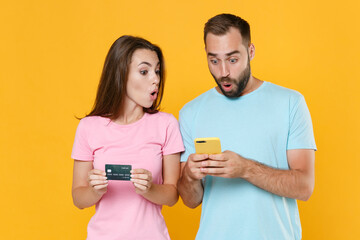 Shocked young couple two friends guy girl in blue pink t-shirts isolated on yellow background studio portrait. People lifestyle concept. Mock up copy space. Using mobile phone, hold credit bank card.