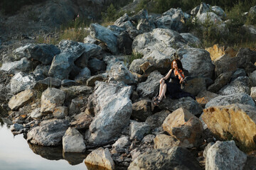 brunette girl with lush curly hair sits on the stones near the lake in summer in a dark blue dress with a violin, professional musician violinist