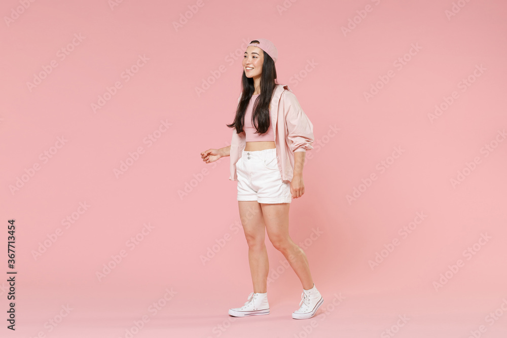 Wall mural Full length portrait of smiling young asian girl in casual clothes, cap isolated on pastel pink background studio portrait. People lifestyle concept. Mock up copy space. Walking going looking aside. - Wall murals