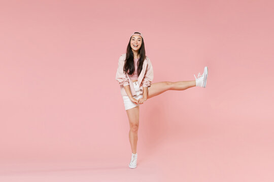 Full length portrait of cheerful young asian girl in casual clothes, cap isolated on pastel pink background studio portrait. People sincere emotions lifestyle concept. Mock up copy space. Rising leg.