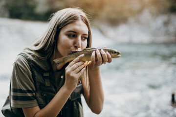 Young adult woman is fishing alone on fast mountain river. The girl holds a live trout and kisses it before releasing it into the river again.