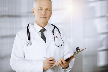 Elderly confident doctor standing straight and using clipboard in sunny clinic. Perfect medical service. Medicine concept