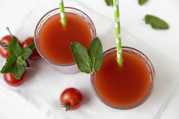 Two glasses with tomato juice stand on a light background, decorated with tubes and mint leaves, next to it are tomato slices