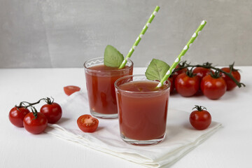 Two glasses with tomato juice stand on a light background, decorated with tubes and mint leaves, next to it are tomato slices